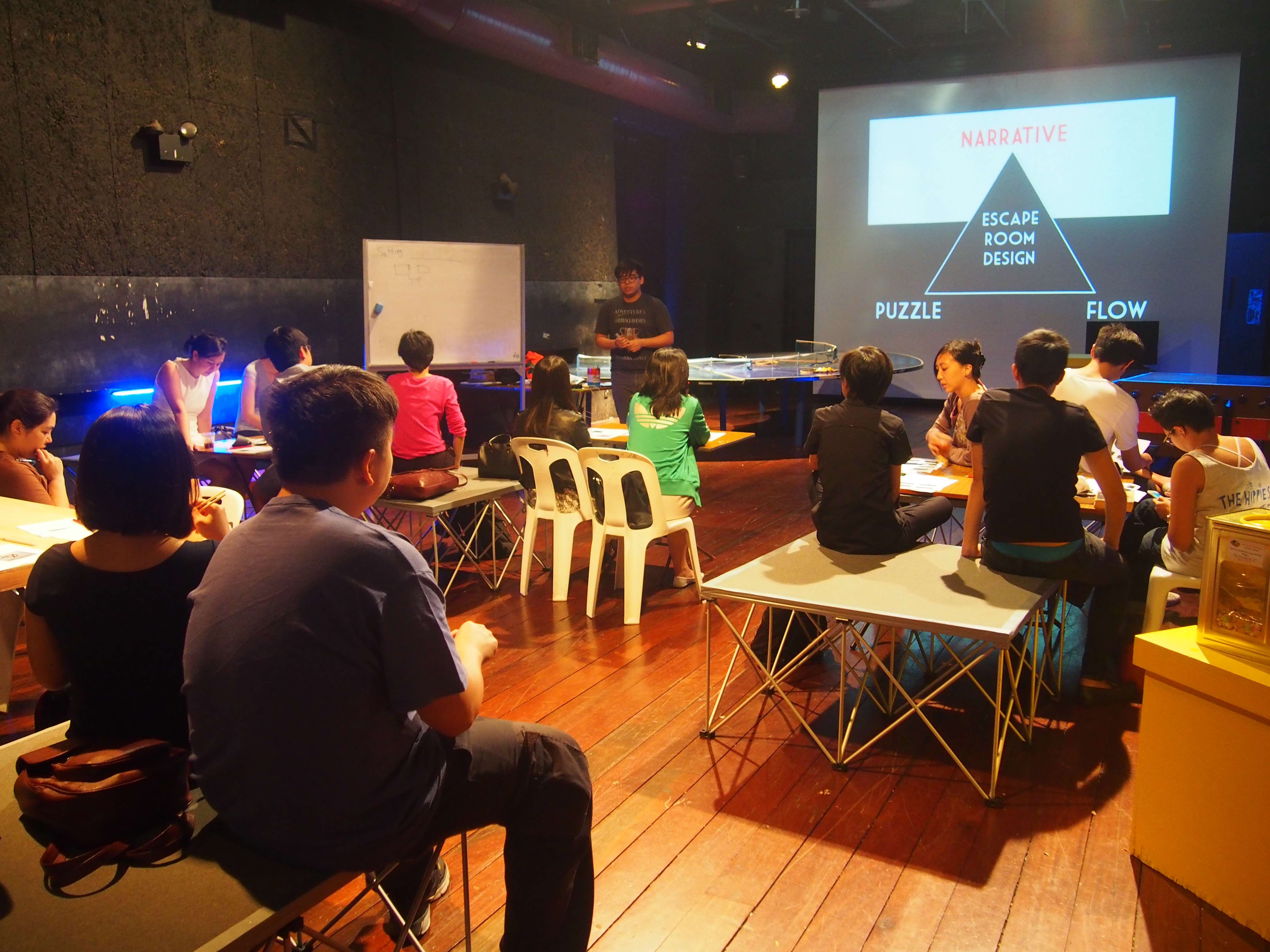 Photograph of a training venue with around ten trainees seated and one trainer who is standing. There are workshop materials such as a whiteboard and projector screen. On the projector screen is a triangular diagram labeled, escape room design, narrative, puzzle, flow.